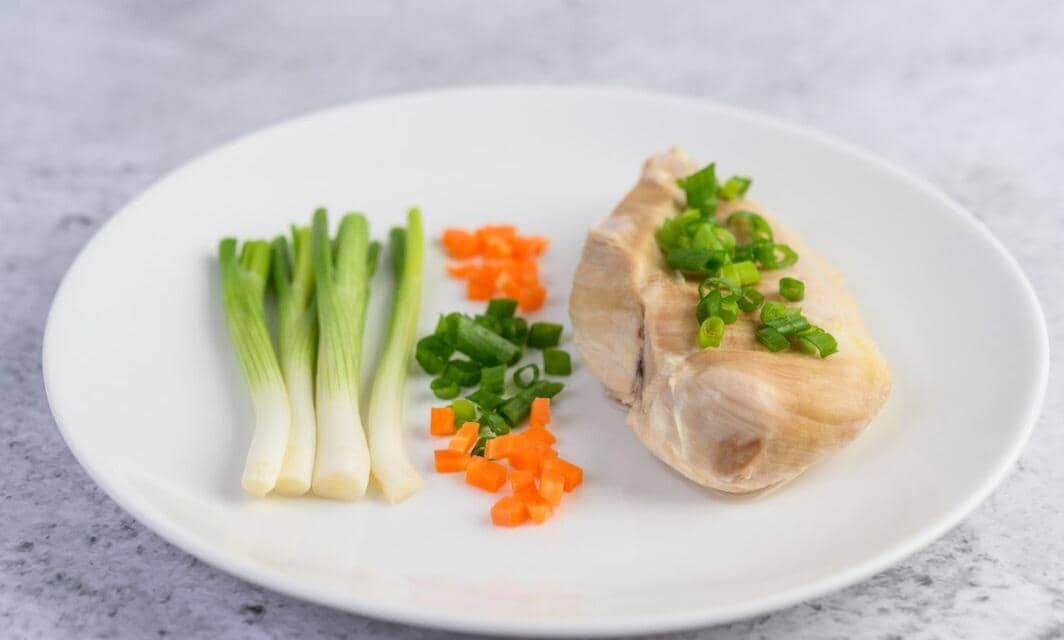 Dada ayam rebus disajikan di atas piring putih dengan potongan wortel, daun bawang cincang, dan bawang daun utuh di sampingnya.