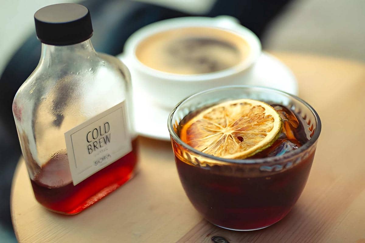 Segelas cold brew dengan irisan lemon di atas meja kayu, disertai botol berlabel cold brew dan secangkir kopi di latar belakang.