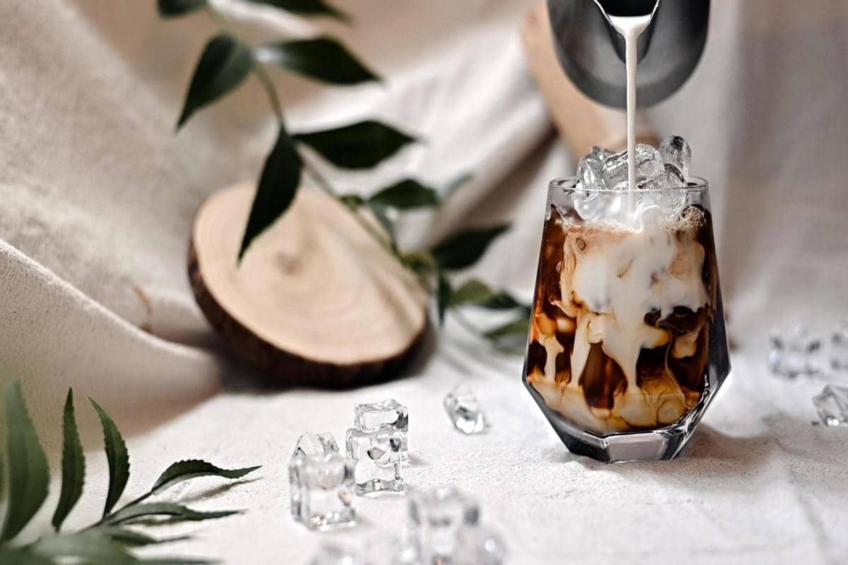 Segelas cold brew dengan susu dituangkan di atas es batu, menciptakan pola berwarna cokelat dan putih yang menarik di atas meja.