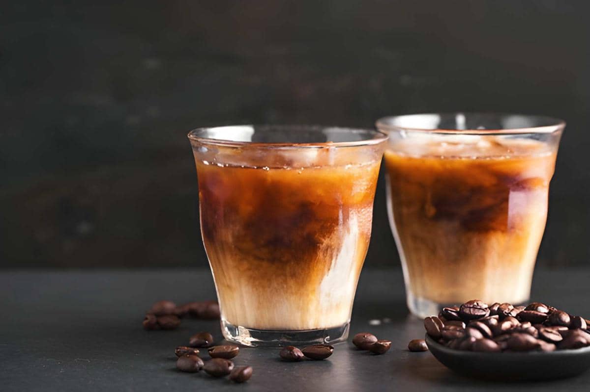 Dua gelas cold brew tonic dengan es batu dan lapisan kopi serta susu di atas meja gelap, dihiasi biji kopi di sekitarnya.
