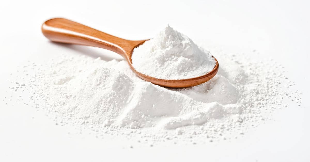Bubuk pengembang (baking powder) berwarna putih dalam mangkuk kecil dengan sendok kayu di atas meja kayu.