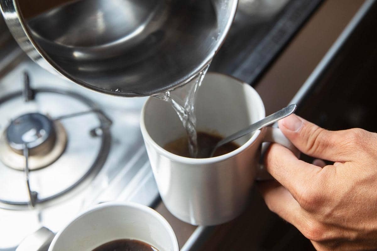 Tangan menuangkan air panas dari panci ke dalam cangkir berisi bubuk kopi di dapur dengan kompor gas di latar belakang.