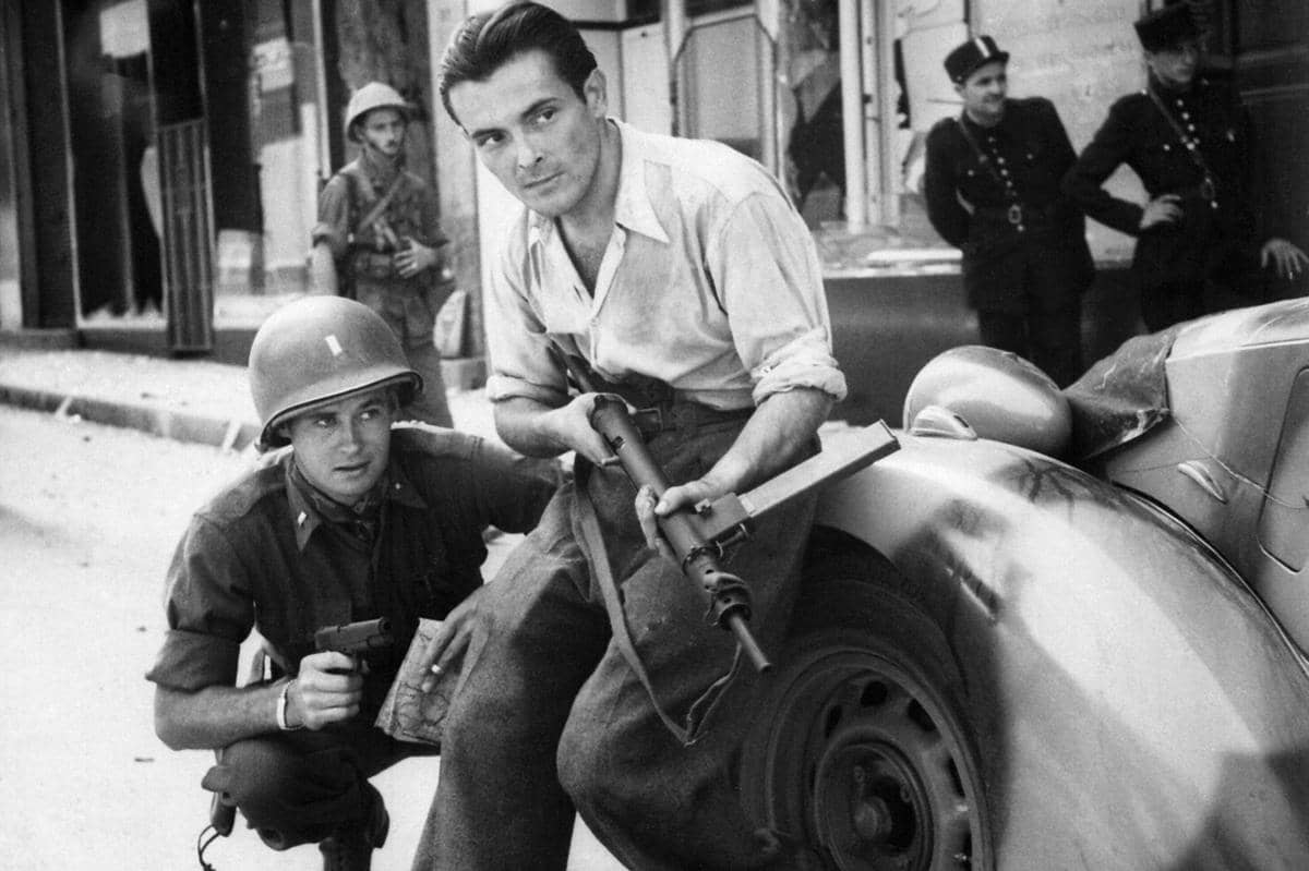 Perwira Amerika dan anggota Perlawanan Prancis bersembunyi di balik sebuah mobil saat terjadi pertempuran di jalanan sebuah kota di Prancis selama Perang Dunia II. 