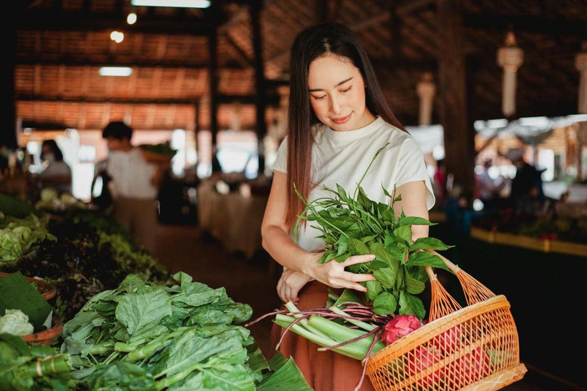 Ilustrasi aktivitas belanja sayur di pasar tradisional, dengan pilihan sayuran segar tersusun di area penjualan.