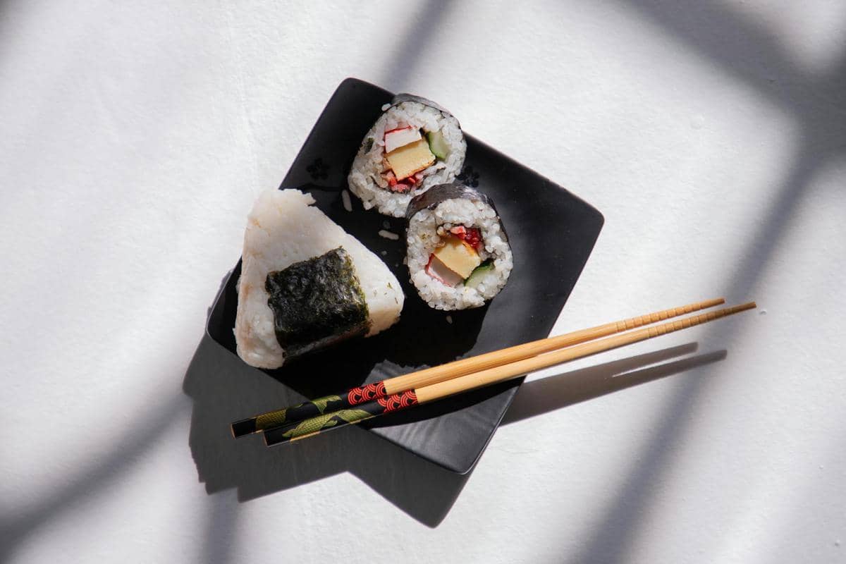 Nasi kepal segitiga dan dua potong sushi gulung disajikan di piring hitam dengan sepasang sumpit kayu di atas meja putih.