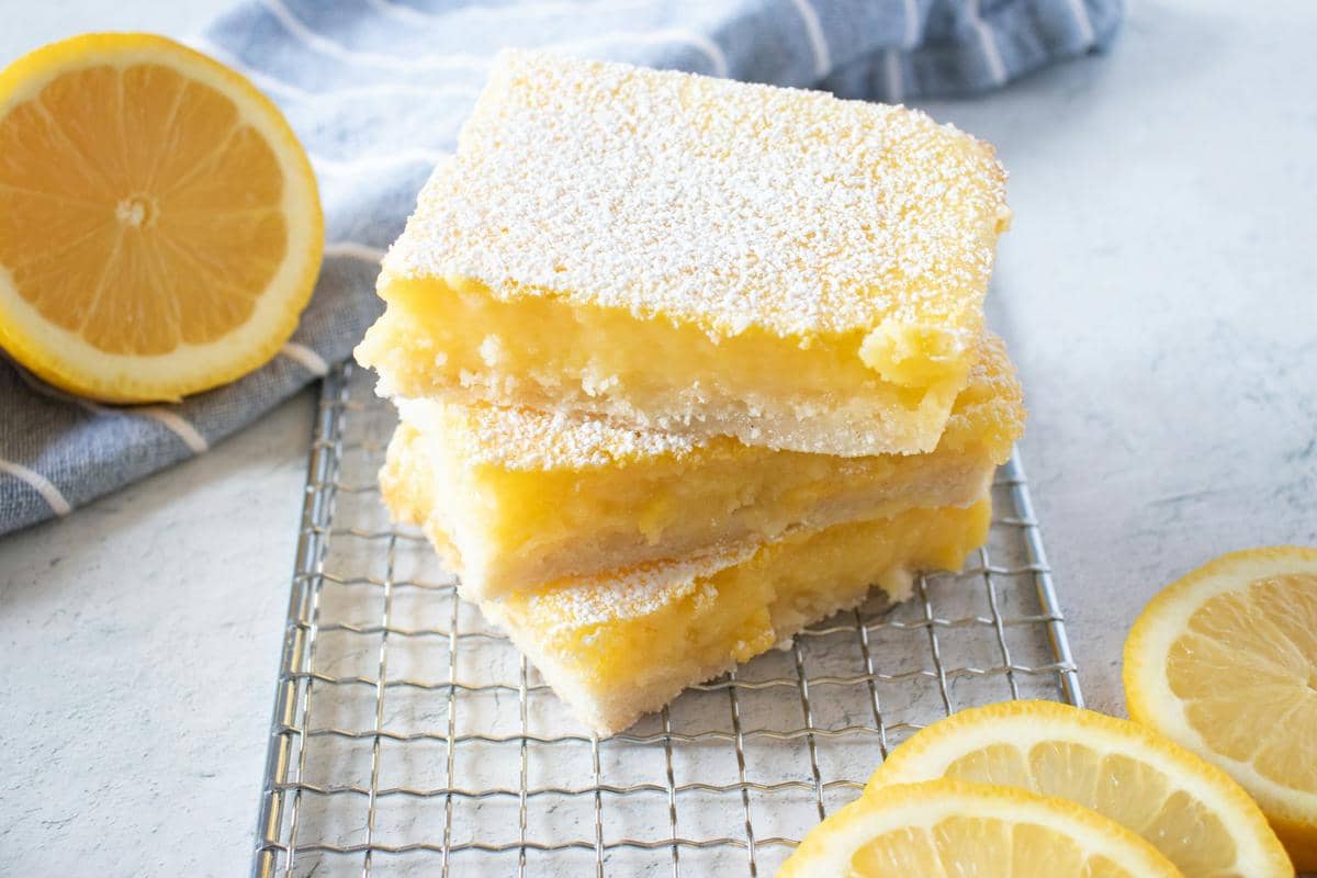 Tiga potong lemon bars berlapis gula halus tersusun di atas rak pendingin dengan irisan lemon segar di sekelilingnya.