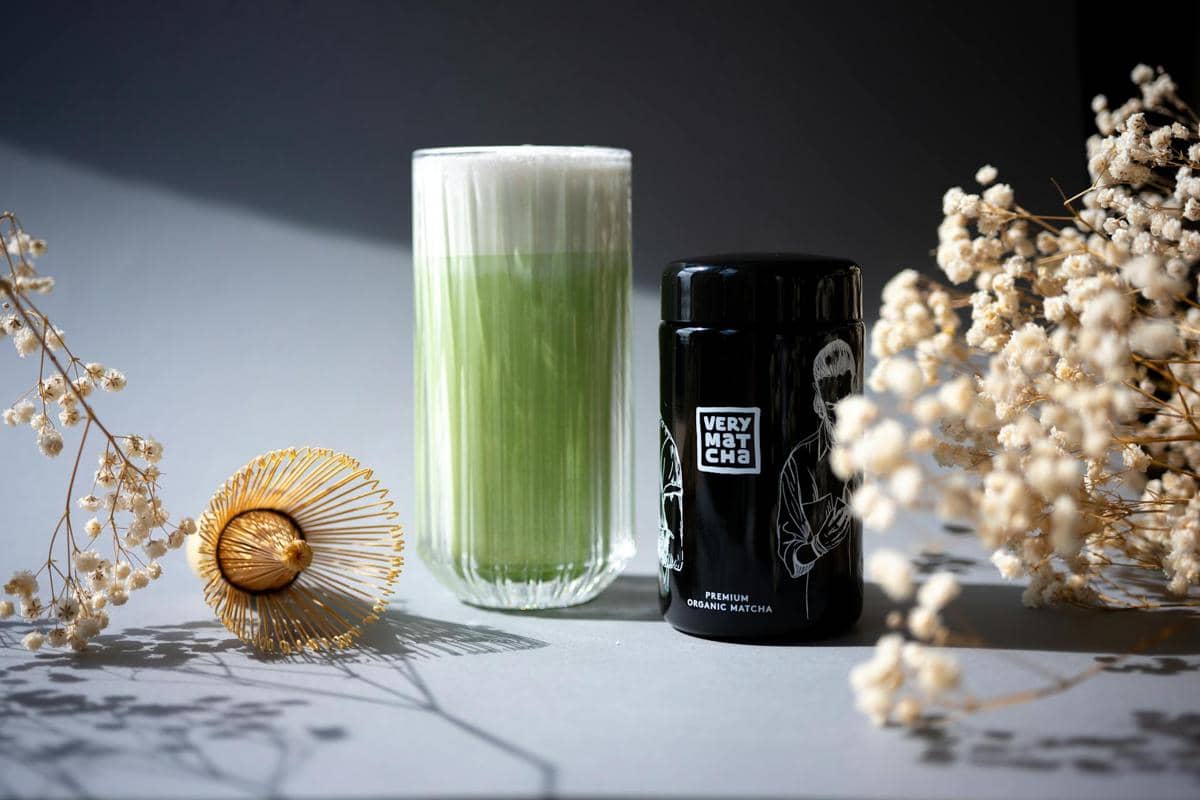 Segelas matcha hijau dengan busa tebal di samping wadah bubuk matcha hitam bertuliskan Very Matcha dan hiasan bunga kering.