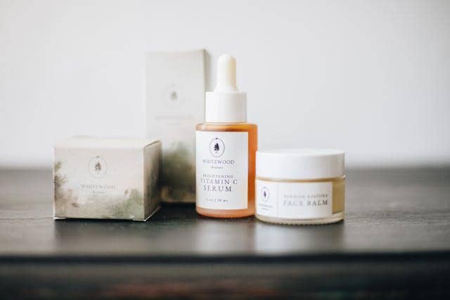 Produk skincare terdiri dari serum vitamin C dan face balm dalam kemasan elegan berwarna netral di atas meja kayu gelap.