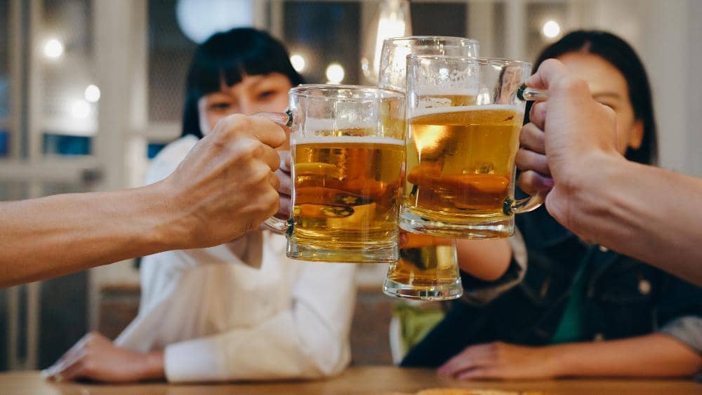 Beberapa orang bersulang dengan gelas berisi minuman berwarna keemasan di meja kayu dalam suasana santai bersama teman.