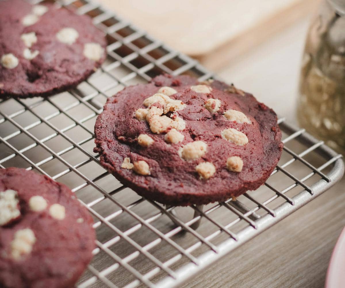 Sejumlah red velvet cookies berwarna merah dengan potongan cokelat putih tersaji di atas alas berwarna terang.