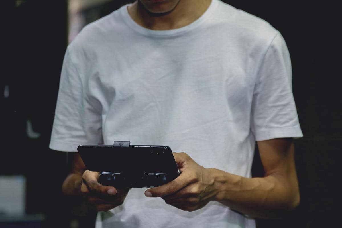 Seorang pria mengenakan kaus putih sedang bermain game di ponsel dengan alat pengendali genggam di lingkungan gelap.