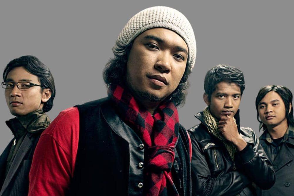 Empat pria berpose bersama di depan latar abu-abu, salah satunya mengenakan topi rajut putih dan syal merah hitam bermotif kotak.