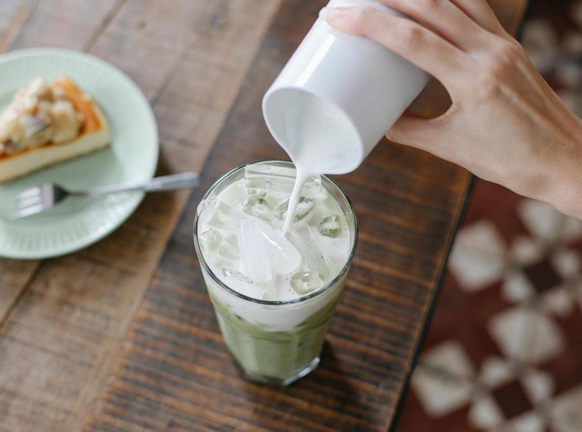 Tangan menuang susu ke dalam segelas minuman hijau berisi es batu di atas meja kayu dengan sepotong kue di piring hijau di sampingnya.