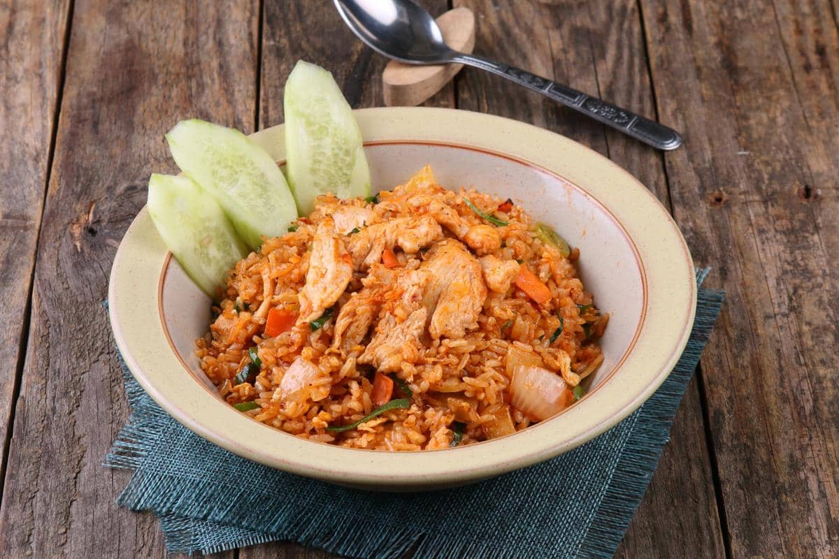 Semangkuk nasi goreng dengan potongan ayam, sayuran, dan irisan mentimun disajikan di atas meja kayu dengan sendok di sampingnya.