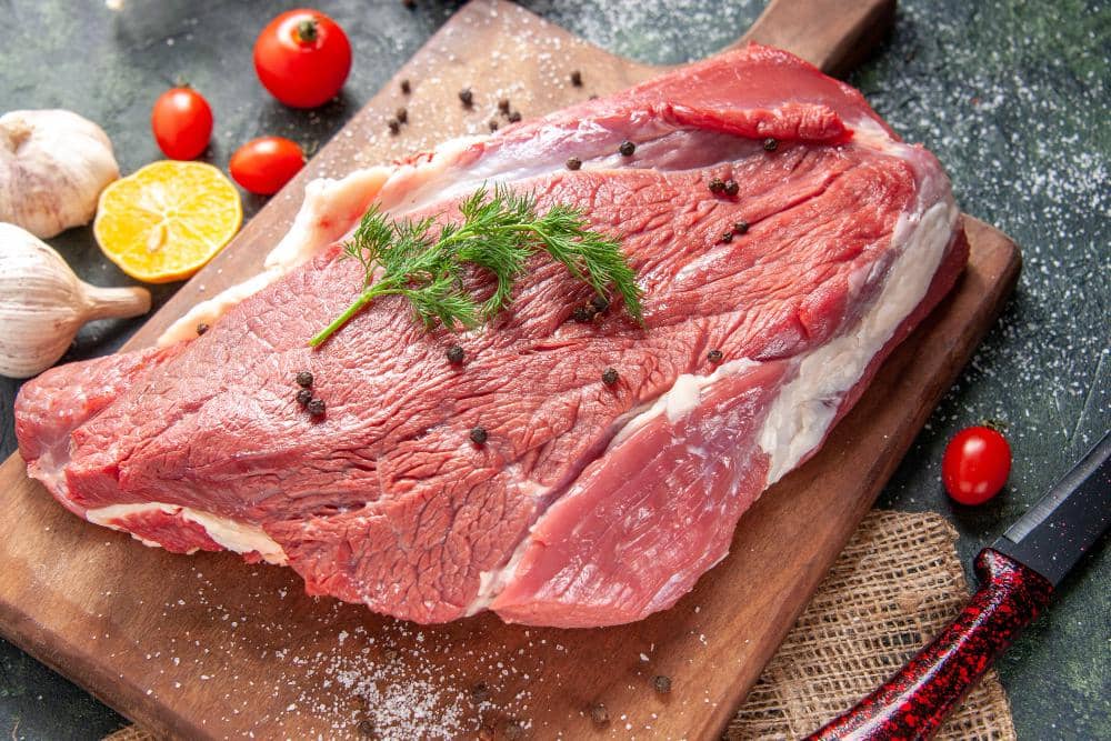 Potongan besar daging sapi mentah di atas talenan kayu dengan taburan lada hitam, bawang putih, tomat, dan irisan jeruk di sekitarnya.