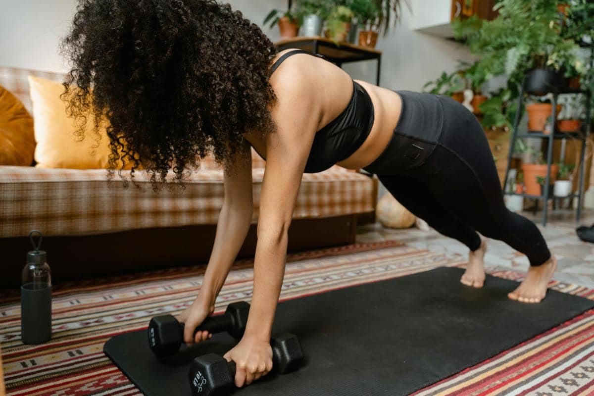 Seorang perempuan berolahraga di rumah dengan posisi plank sambil memegang dua dumbbell di atas matras hitam di ruang tamu.