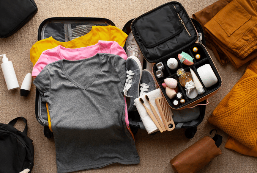 Koper terbuka berisi pakaian berwarna abu-abu, kuning, dan merah muda, sepatu, serta perlengkapan skincare dan alat mandi untuk traveling.