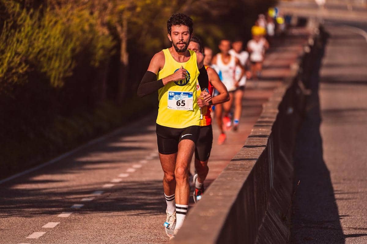 Pelari mengenakan kaus kuning dan nomor dada 65 berlari di depan peserta lain dalam lomba marathon di jalan raya.