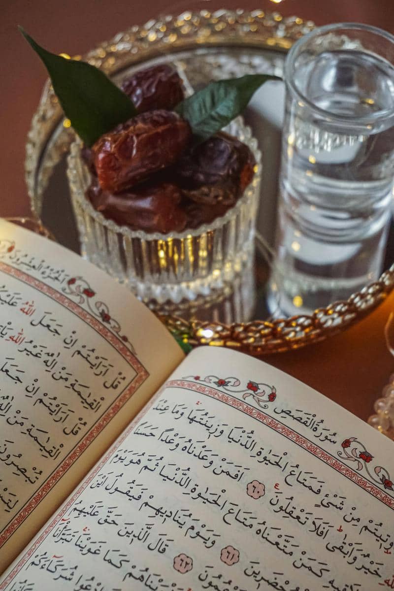 Foto Al-Qur’an terbuka dengan kurma dan segelas air di atas nampan, menggambarkan suasana berbuka puasa dan persiapan ibadah Ramadan.