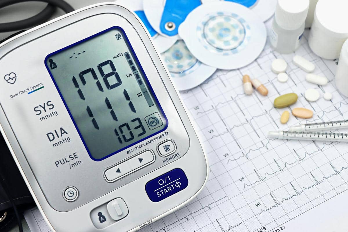 Alat pengukur tekanan darah digital menunjukkan hasil tinggi di atas kertas EKG dengan beberapa obat dan jarum suntik di sekitarnya.