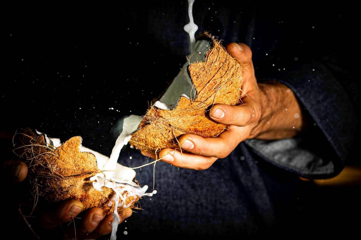 Tangan memegang kelapa yang dibelah dua dengan santan mengalir keluar, latar belakang gelap menonjolkan tekstur kelapa.