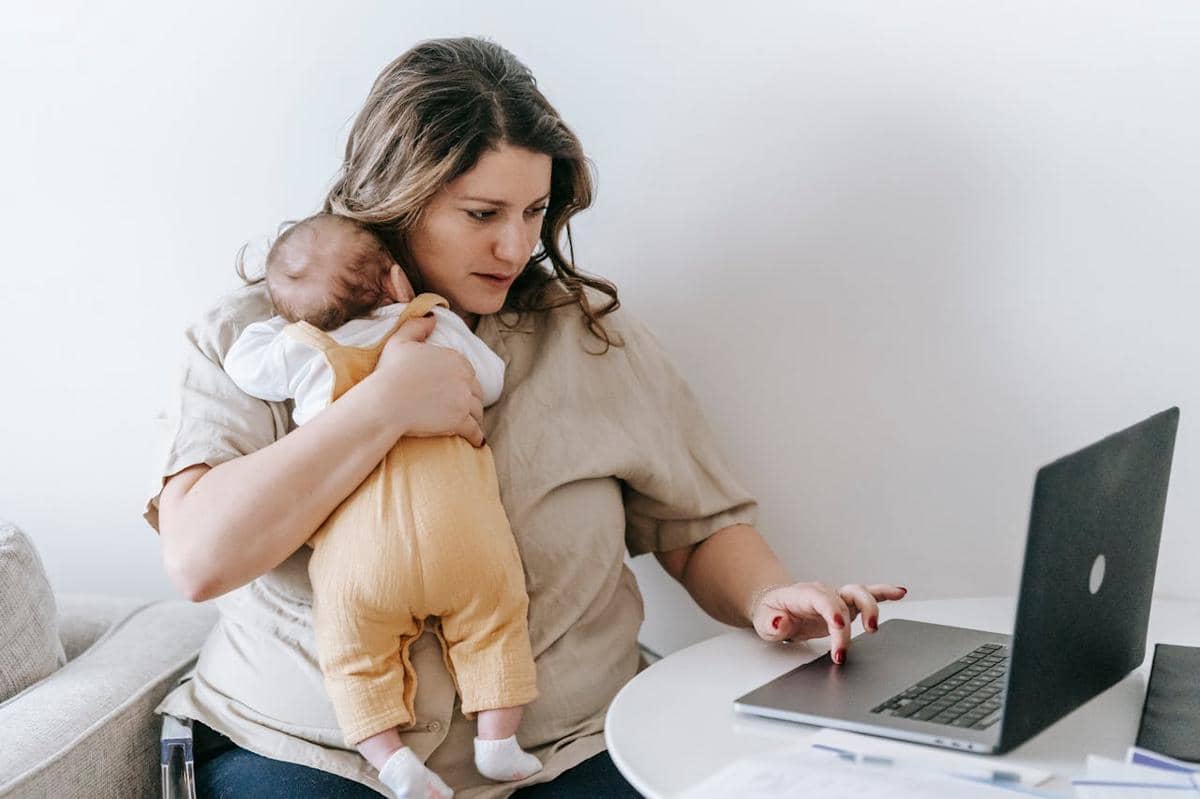 Seorang ibu bekerja menggendong bayi sambil menggunakan laptop di rumah, menggambarkan keseimbangan pekerjaan dan pengasuhan.