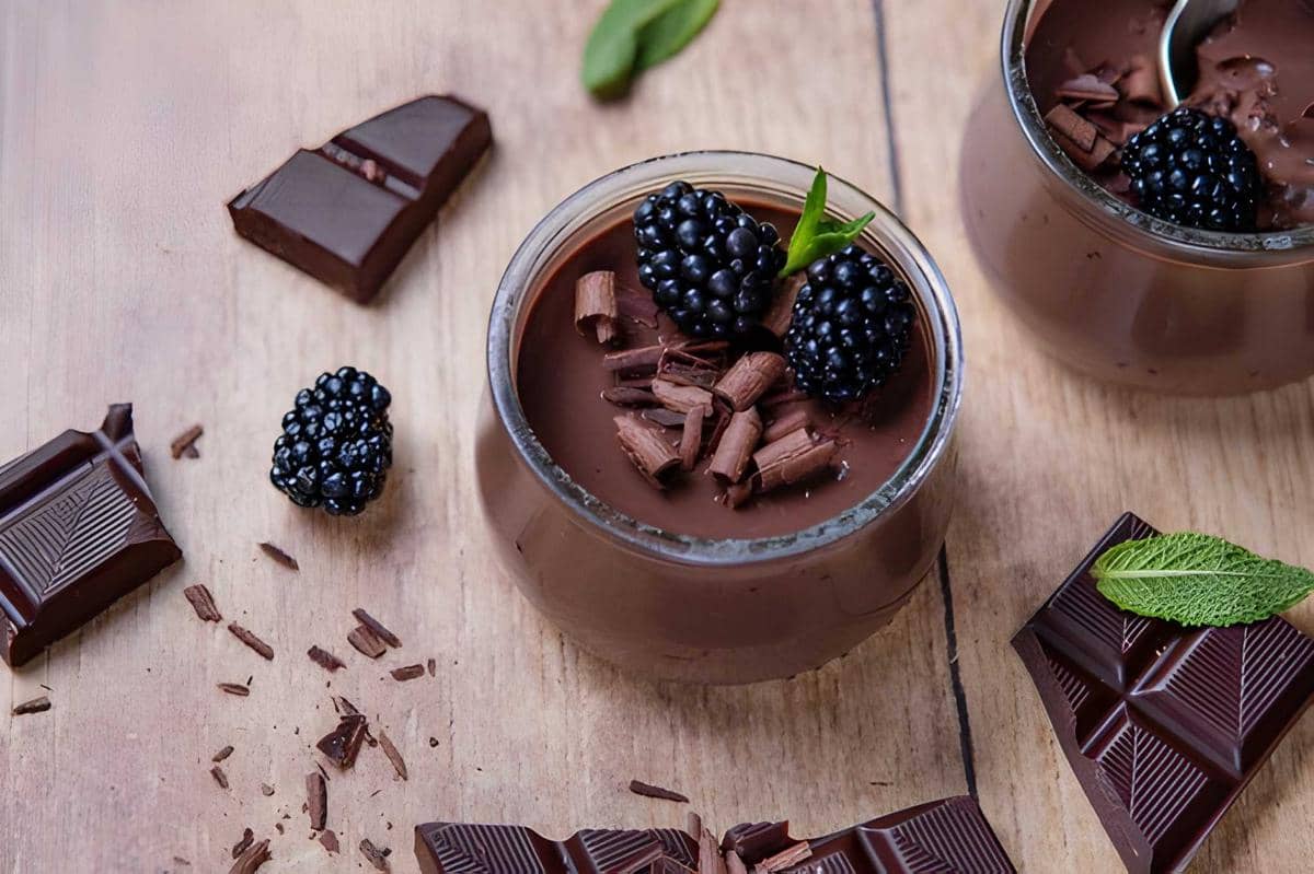 Puding cokelat dalam gelas kecil dihiasi potongan cokelat dan buah blackberry segar di atas meja kayu dengan potongan cokelat di sekitarnya.