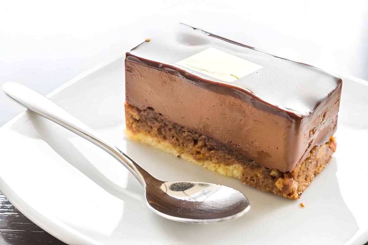 Sepotong cheesecake cokelat tanpa panggang dengan lapisan krim lembut dan dasar biskuit, disajikan di piring putih bersama sendok perak.