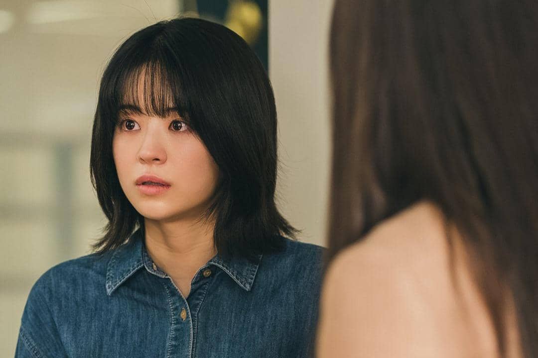 Seorang wanita berambut pendek mengenakan kemeja denim tampak terkejut saat berbicara dengan seseorang di depannya.