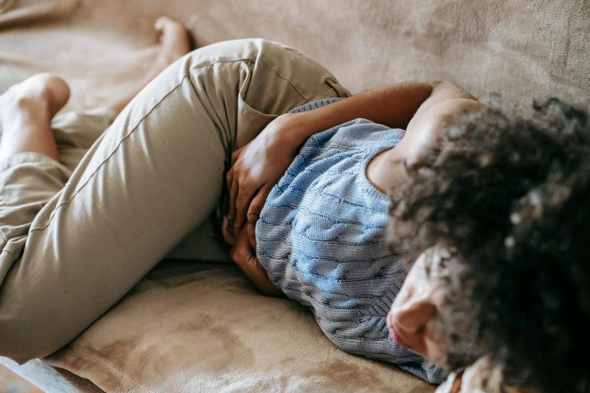 Seorang perempuan berbaring di sofa sambil memegangi perutnya, tampak kesakitan akibat nyeri perut yang dialaminya.