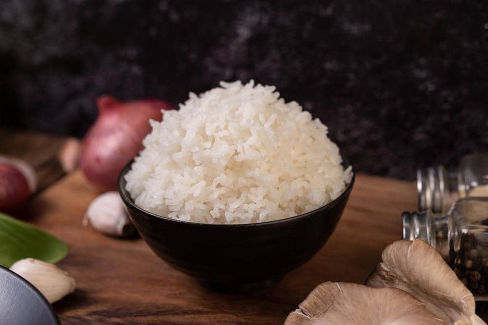 Semangkuk nasi putih pulen dalam wadah hitam dengan butiran beras tersebar di atas meja kayu sebagai ilustrasi makanan pokok.