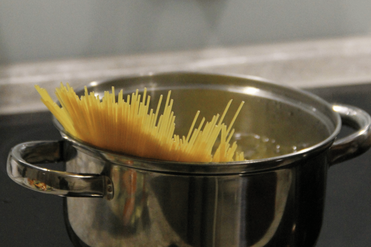 Panci berisi air mendidih di atas kompor dengan spageti kering yang mulai dimasukkan ke dalam air untuk direbus.