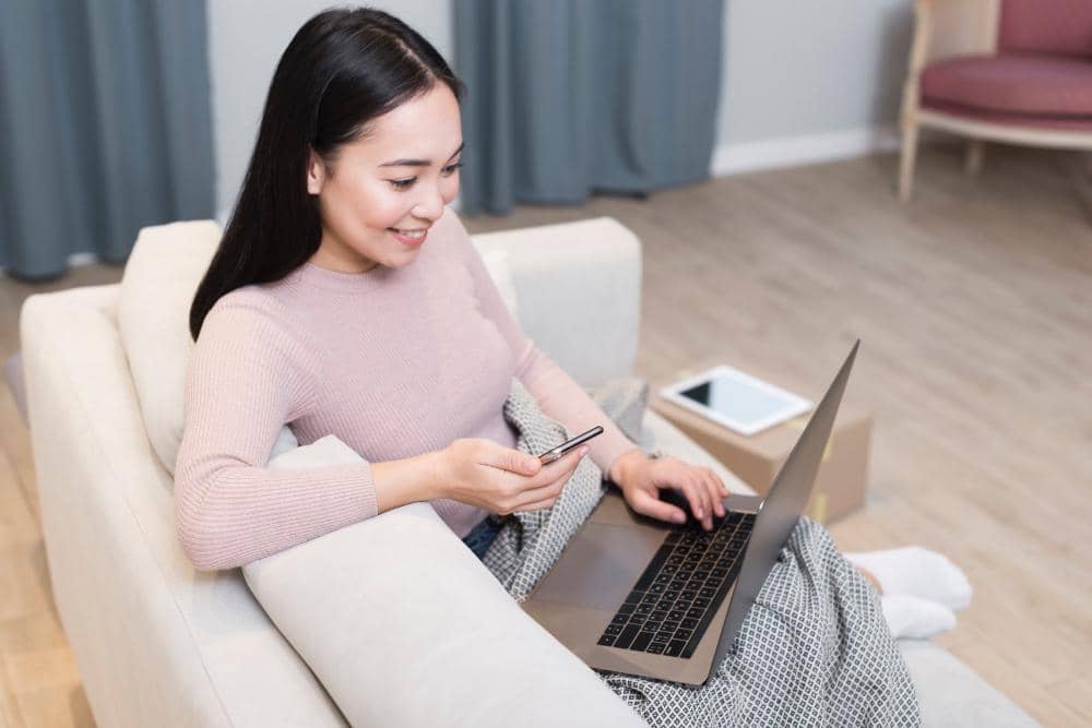 Seorang wanita duduk di sofa sambil bekerja dengan laptop dan memegang ponsel, menggambarkan aktivitas admin media sosial.