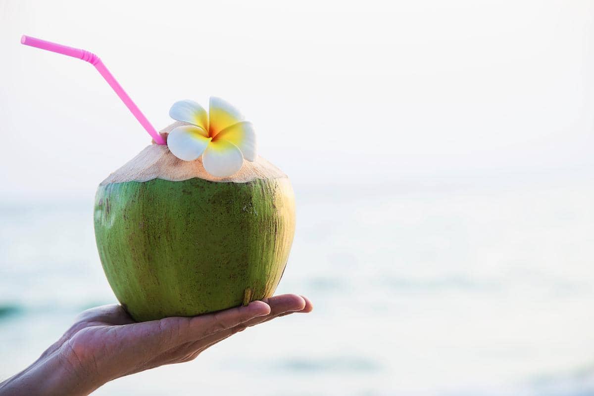 Tangan memegang es kelapa muda dengan sedotan dan bunga kamboja di pantai berpasir, berlatar ombak laut.