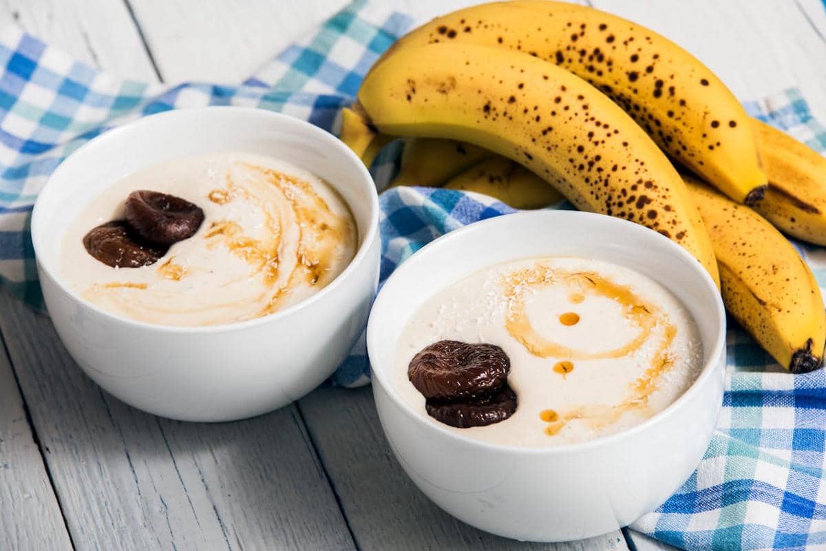 Dua mangkuk yogurt dengan topping buah kering dan madu di atas meja kayu putih, disertai beberapa pisang matang di latar belakang.