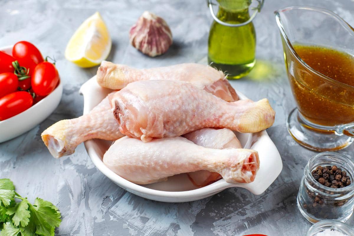 Beberapa potong paha ayam mentah di atas piring putih dengan bahan masakan seperti tomat, bawang putih, lemon, dan minyak zaitun di sekitarnya.
