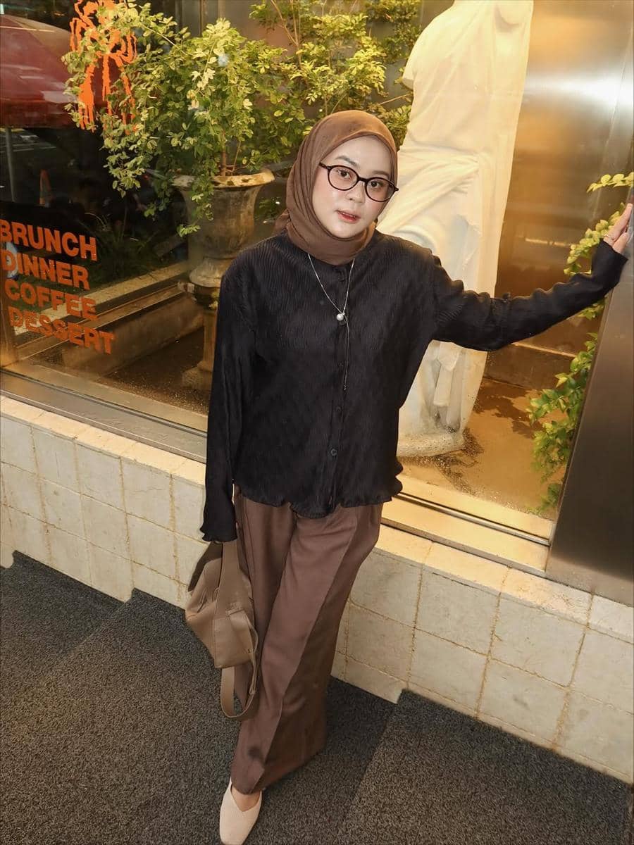 Seorang wanita berhijab cokelat mengenakan kemeja hitam dan celana panjang cokelat, berpose di depan jendela kaca dengan tanaman hias di belakangnya.