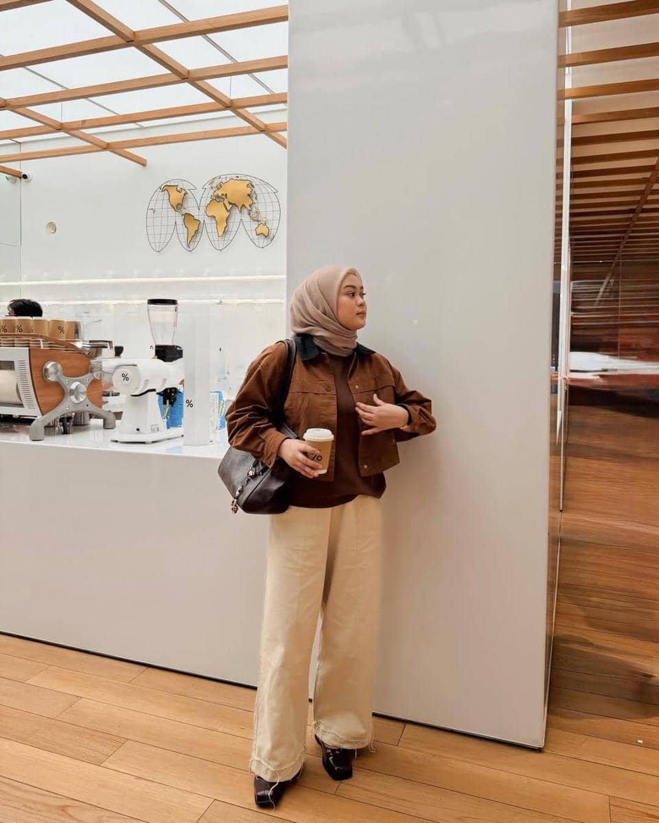 Seorang wanita berhijab mengenakan jaket cokelat dan celana krem sedang memegang kopi di sebuah kafe modern dengan interior kayu.