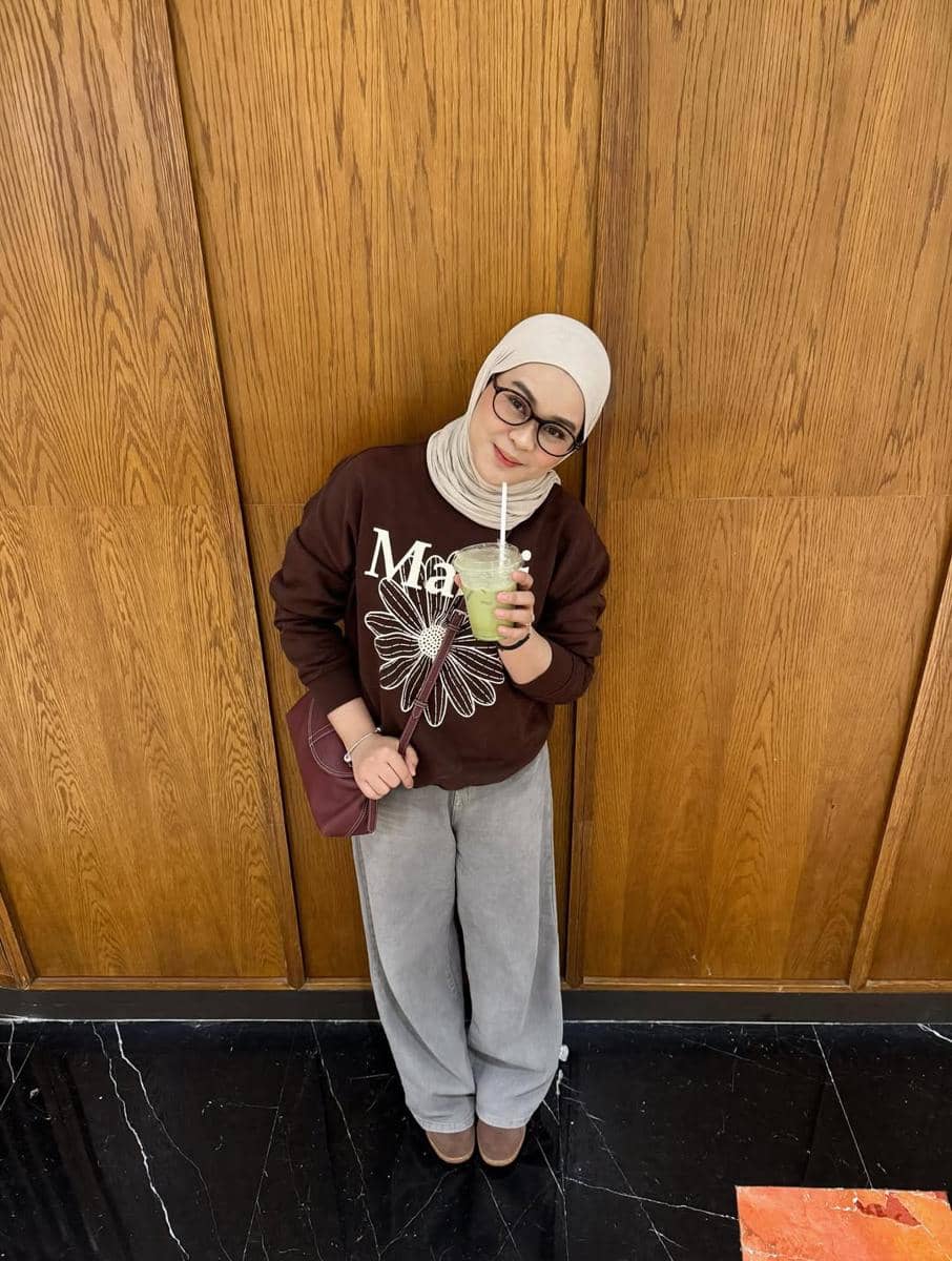 Seorang wanita berhijab mengenakan sweater cokelat bergambar bunga, celana abu-abu longgar, dan membawa minuman hijau di depan dinding kayu.