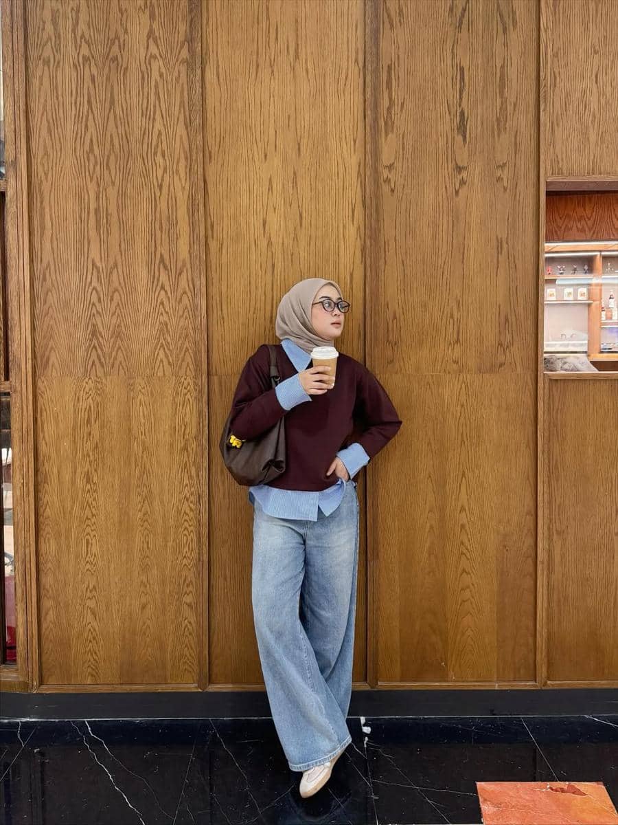 Seorang wanita berhijab mengenakan sweater marun, kemeja biru, dan celana jeans longgar sambil memegang kopi di depan dinding kayu.