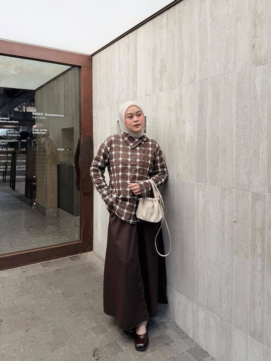 Seorang wanita berhijab mengenakan sweater motif kotak cokelat dan rok panjang cokelat tua sambil memegang tas krem di depan dinding beton.