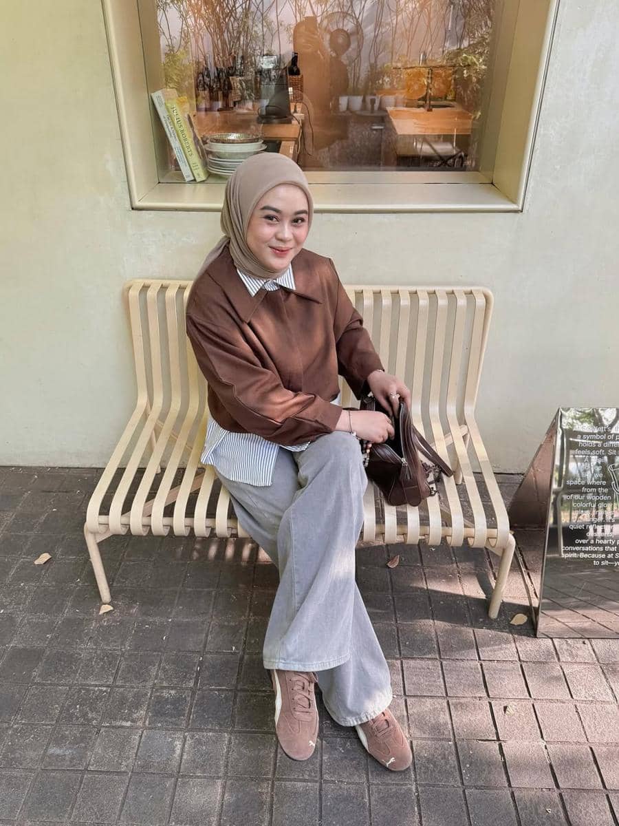 Seorang wanita berhijab duduk di bangku luar ruangan mengenakan sweater cokelat, kemeja bergaris, celana abu-abu, dan sepatu cokelat.