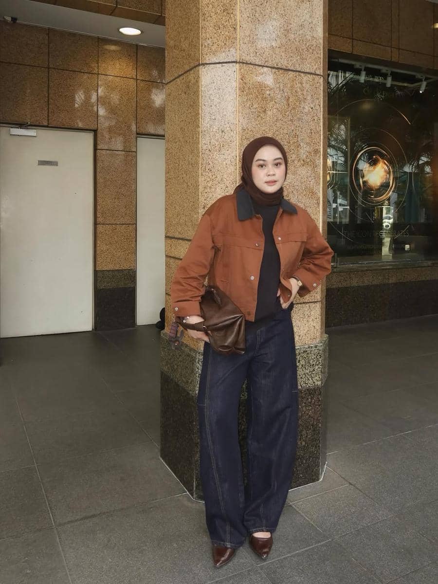 Seorang wanita berhijab mengenakan jaket cokelat, atasan hitam, dan celana jeans longgar sambil berpose di depan gedung bertembok granit.