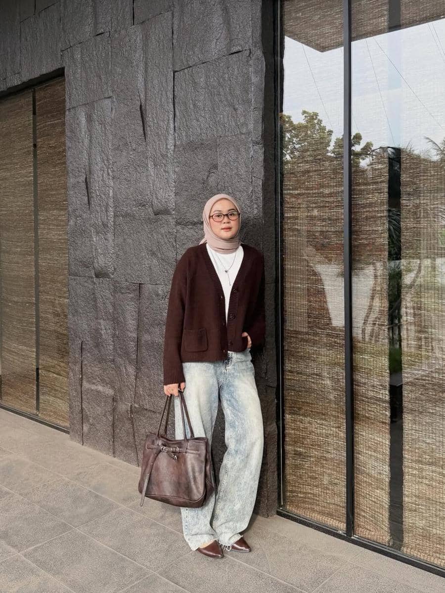 Seorang wanita berhijab mengenakan kardigan cokelat tua, celana jeans longgar, dan membawa tas kulit cokelat di depan dinding batu abu-abu.