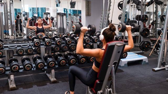 Seorang wanita berolahraga di gym sambil mengangkat dumbbell di depan cermin, mengenakan pakaian olahraga hitam dan headphone.