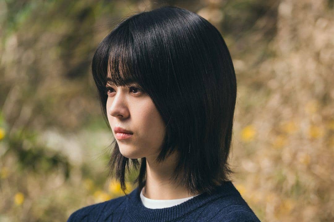 Seorang perempuan berambut pendek hitam mengenakan sweater biru tampak menatap ke arah samping di luar ruangan dengan latar alam.