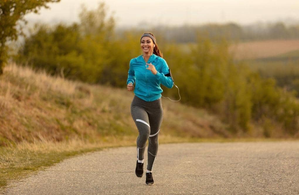 Seorang perempuan berlari mengenakan jaket biru dan celana olahraga abu-abu sambil tersenyum menikmati udara segar.