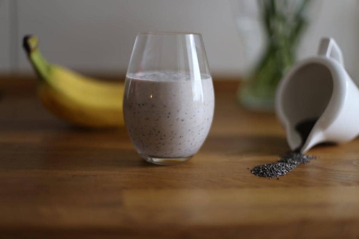 Segelas smoothie pisang berwarna krem dengan biji chia di atas meja kayu, di samping pisang dan wadah kecil berisi biji chia tumpah.