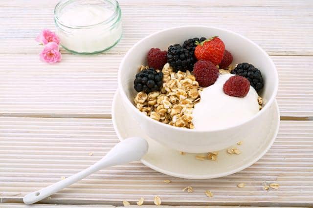 Semangkuk overnight oat dengan yogurt, granola, dan buah beri segar seperti stroberi, raspberry, serta blackberry di atas meja kayu putih.