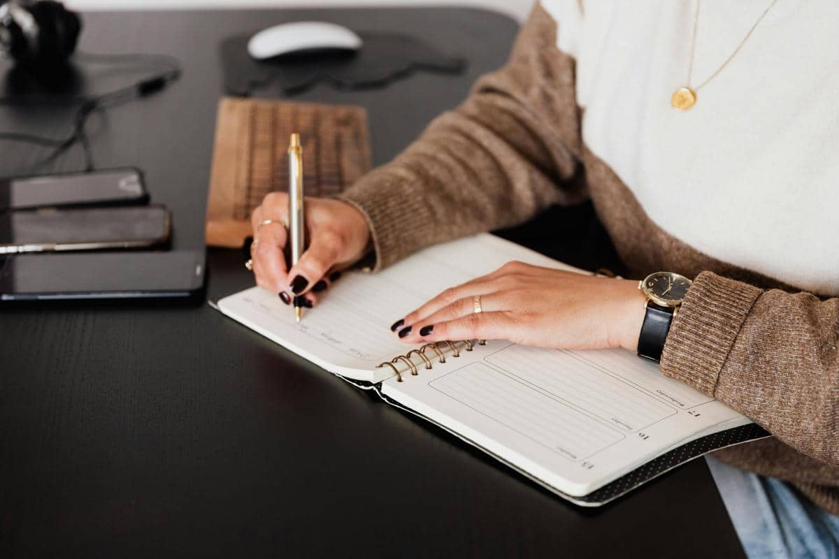 Seseorang menulis jadwal di buku agenda di atas meja kerja hitam dengan ponsel dan laptop di sekitarnya.